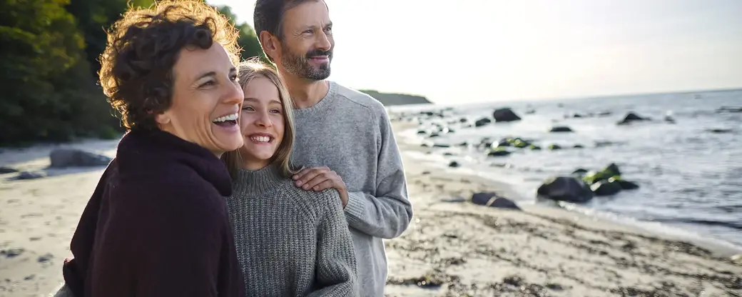 A group of three people—two women and one man—are standing together on a beach, smiling and enjoying each other’s company. They are dressed in warm sweaters, with the ocean and rocks in the background under a bright sky.
