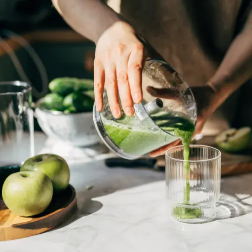 A hand pours a vibrant green juice from a blender into a clear glass. Fresh green apples and cucumbers are visible on the countertop, highlighting the healthy ingredients used for the drink. The scene is bright and fresh, emphasizing a focus on nutritious preparation.