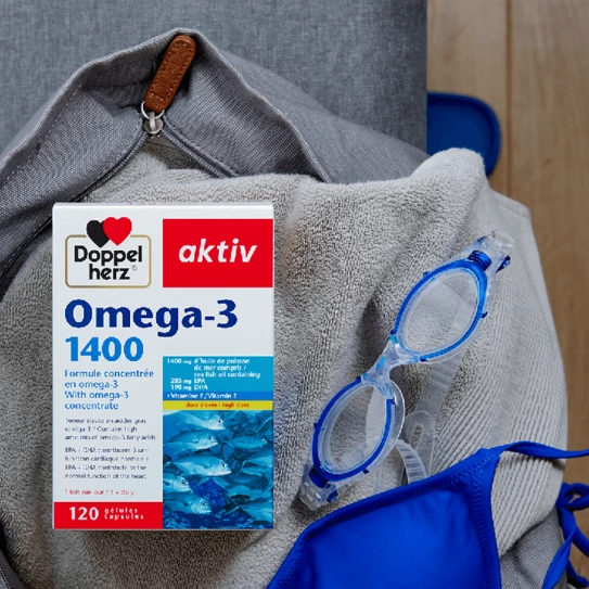 A box of Doppelherz aktiv Omega-3 dietary supplement is placed on a soft gray towel. The box highlights it contains 1400 mg of Omega-3 from fish oil. Next to it are a pair of blue swimming goggles and a blue garment, suggesting a swimming theme.