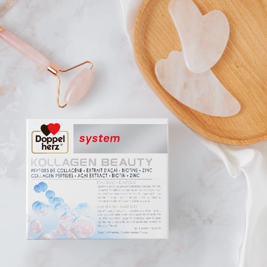 A box of Doppelherz Kollagen Beauty sits on a light surface, displaying benefits like collagen peptides, biotin, and zinc. Nearby, a wooden plate holds two heart-shaped, translucent facial tools, alongside a facial roller. The setting emphasizes beauty and skincare.