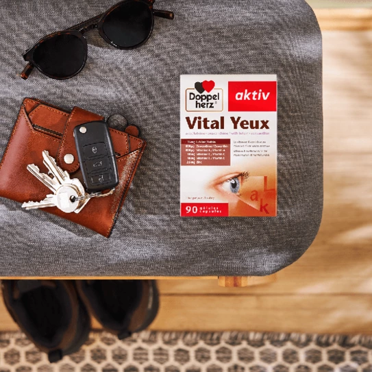 A brown wallet with keys and a black car remote lies on a textured surface. Next to it is a box labeled "Doppelherz aktiv Vital Yeux," indicating it contains 90 capsules for eye health. Sunglasses rest nearby, suggesting a casual, outdoor lifestyle.
