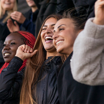 Three women are smiling and laughing together, expressing joy and excitement. The woman on the left has dark skin and red braided hair, while the others have light skin and long hair. They are wearing casual jackets and seem to be in a lively atmosphere, possibly at an event or celebration.